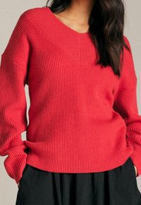 Czerwony sweter w prążki z dekoltem w serek i d�ługimi rękawami, z fakturowanym wzorem dzianiny i luźnym krojem. Połączony z czarnym ubraniem pod spodem.