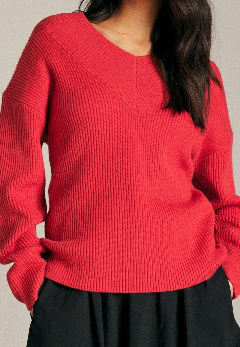 Czerwony sweter w prążki z dekoltem w serek i długimi rękawami, z fakturowanym wzorem dzianiny i luźnym krojem. Połączony z czarnym ubraniem pod spodem.