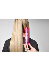 En rosa sprayflaska märkt "Redken" hålls nära rakt, långt blont hår, mot en neutral bakgrund med tydlig textur.