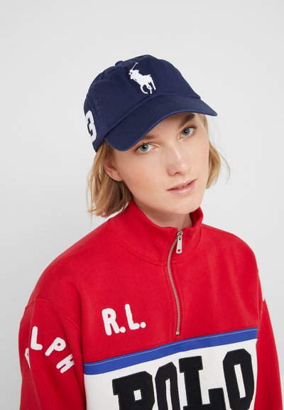 Ναυτικό καπέλο μπέιζμπολ με λευκό κεντημένο λογότυπο παίκτη polo και αριθμό 3. Κόκκινη ζακέτα με φερμουάρ με λευκές και μπλε ρίγες, που φέρει το κείμενο "POLO".
