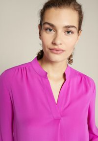 Rosa Bluse mit V-Ausschnitt, weichem Stoff und dezenten Falten an den Schultern. Nahaufnahme, die sich auf den Kragen und den oberen Torso konzentriert.