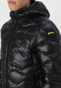 Giacca a vento nera con design trapuntato, texture lucida, chiusura con zip, tasca frontale e accent in giallo con logo sulla manica.