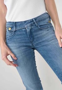 Mujer con jeans pitillo de talle alto azul con detalle de botones dorados y top blanco sin mangas de canalé, con las manos descansando a los lados.