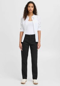 Pantaloni neri in cotone abbinati a una camicia bianca con bottoni sopra una canotta bianca, indossati con sneakers color crema. Design a gamba dritta, dettagli minimi.