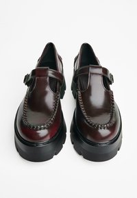 Burgunderfarbene Lederschuhe mit glänzender Oberfläche, die ein gesticktes Design, einen Riemen mit Schnalle und eine dicke schwarze Plateausohle aufweisen.