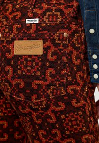 Wrangler corduroybyxor med ett flerfärgat, geometriskt mönster i rött, orange och brunt med en synlig märke-patch och metallknapp-accenter.