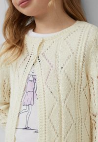 Cardigan en maille de couleur beige crémeux avec un motif en diamant, doté d'une fermeture à boutons et d'une texture ajourée. En dessous, un t-shirt blanc avec un design de dessin animé.