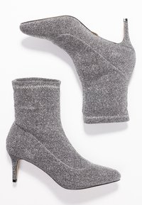 Bottines à paillettes argentées avec un design ajusté et sans couture, bout pointu et talon de 3 pouces. Présente des coutures contrastantes le long de l'ouverture.