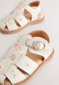 Sandales en cuir blanc avec broderie florale en rose et jaune, dotées d'une fermeture à boucle, d'un design ouvert et d'une semelle naturelle imitant le bois.