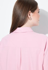 Camicia color rosa chiaro con colletto, caratterizzata da una piega posteriore. Texture liscia con vestibilità comoda, adatta per un abbigliamento casual.