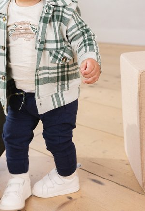 Małe dziecko w białych tenisówkach, granatowych spodniach, beżowej koszuli z nadrukiem misia oraz zielonej kratkowanej kurtce stoi na drewnianej podłodze blisko beżowej sofy.