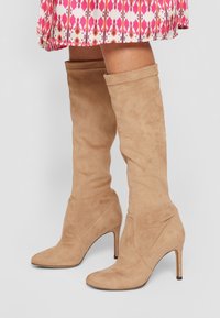 Femme portant des bottes à talons haut en suède beige jusqu'au genou, associées à une jupe à motifs rose et blanche, se tenant sur un fond clair.