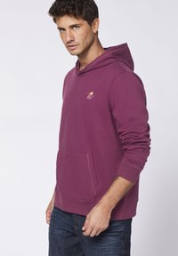 Maroon hooded sweatshirt with a kangaroo pocket and ribbed cuffs. Features a small embroidered logo on the chest. Soft cotton blend material.