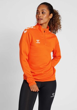 Frau mit lockigem Haar, die ein orangenes Langarm-Quarter-Zip-Oberteil und schwarze Leggings trägt, beide mit weißem hummel-Logo und Chevron-Streifen.