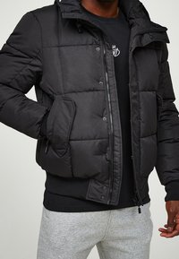 Sort dynejakke med lynlåslukning og trykknapper, med hætte, tekstureret stof og sidelommer. Kombineret med en sort skjorte og grå joggebukser.