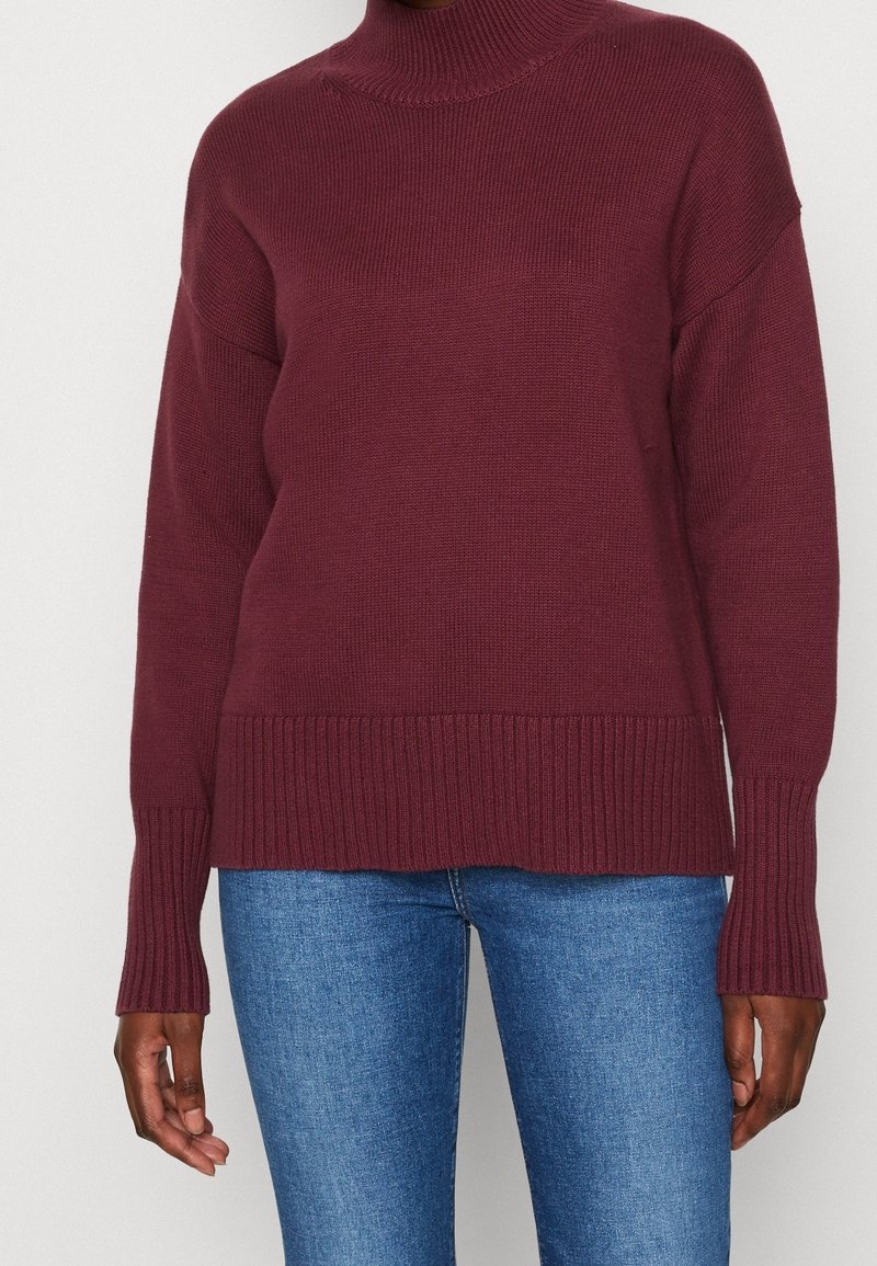 Pull en maille bordeaux avec un col montant, des poignets et un ourlet côtelés ; associé à un jean en denim bleu offrant une coupe décontractée.