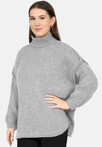 Vrouw met lang bruin haar, gouden oorringen en een grijze oversized gebreide coltrui, poserend met één hand op de heup.