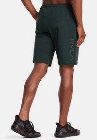 Shorts de sport vert foncé avec un cordon de serrage à la taille et le logo RVCA en gris clair sur le côté, associés à des baskets en mesh noires.