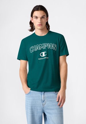 Camiseta de algodón color azul verdoso con el logo "CHAMPION" en impresión blanca. Mangas cortas, cuello redondo, corte relajado. Combinada con vaqueros de mezclilla azul claro.
