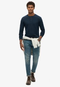 Marinefarbener Langarm-Henley-Pullover mit drei Knöpfen, helle Jeans und braune Schnürstiefel. Hellgrauer Pullover an der Taille gebunden.
