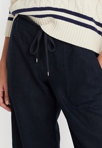 Tmavě modré kalhoty z lnu se dvěma předními kapsami, stahovacím páskem v pase a kovovými koncovkami na tkaničkách. Texturovaná látka s volným střihem.