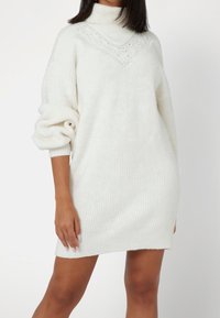 Abito maglione bianco con collo alto, texture a costine e dettagli a trecce sulla scollatura. Maniche ampie e oversize.