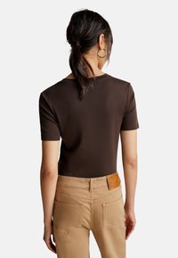 Vrouw met donker haar in een lage paardenstaart, gekleed in een bruin shirt met korte mouwen en beige broek, van achteren afgebeeld op een witte achtergrond.