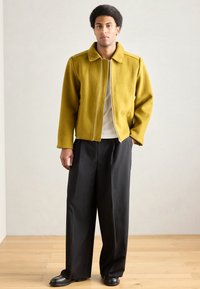 Jakna iz flisa gorčično rumene barve z ravnim krojem, z zadrgo in vezenim ovratnikom, združena z ohlapnimi črnimi hlačami in črnimi čevlji.