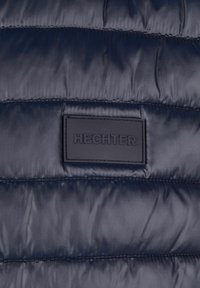 Marineblauer Steppjacke mit gestepptem Muster, glattem Stoff und einem rechteckigen schwarzen Etikett, das das Wort "HECHTER" geprägt hat.