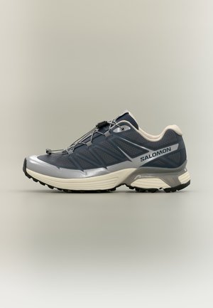 Chaussure de sport Salomon bleu foncé et grise avec semelle blanche, vue latérale montrant un tissu mesh et un système de laçage rapide sur fond neutre.