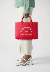 Raudona rankinė su logotipo tekstu, pagaminta iš audinio, su trumpomis rankenomis. Dėvima su sidabriniais kulnais ir kreminėmis kelnėmis. Minimalistinis dizainas.