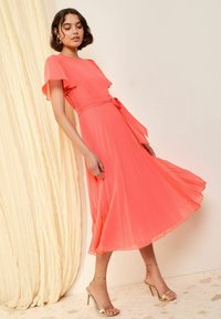 Korallefarbenes midi Kleid mit Faltenrock, Flügelärmeln, einem Stoffband in der Taille und einem schwingenden Rock. Kombiniert mit goldenen, schnüren Sandalen mit Absatz.