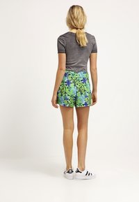 Shorts i livlig grön färg med blå och lila bladmönster, parat med en grå t-shirt. Vita skor med svarta ränder fulländar looken.