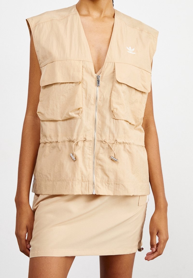 Beige ärmlös väst i lätt tyg, med två framfickor, dragkedjestängning och justerbara dragsnören i fållen.