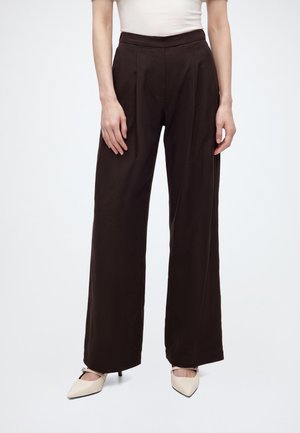 Mujer vistiendo pantalones de pierna ancha de tiro alto en color marrón oscuro, combinados con tacones blancos de punta afilada sobre un fondo blanco liso.