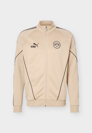 Beige Sportjacke mit Reißverschluss, schwarzer Paspelierung, Puma-Logo und "KING"-Text auf der linken Brust, BVB 09 Emblem auf der rechten Brust, gerippte Bündchen und Saum.