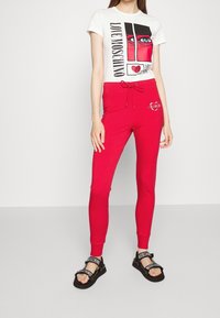 Biała koszulka z graficznym nadrukiem przedstawiającym oczy i serca, zestawiona z czerwonymi dresami z regulowanym paskiem i detalami logo, noszona z czarnymi sandałami.