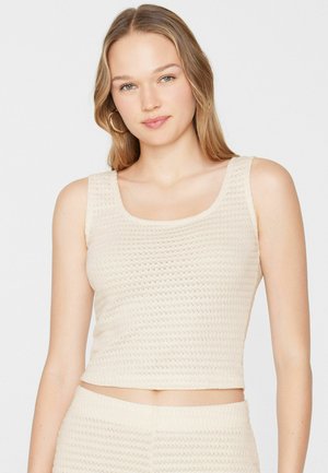 Jeune femme aux longs cheveux blonds portant un crop top en maille crème sans manches et un bas assorti taille haute, debout devant un fond uni.