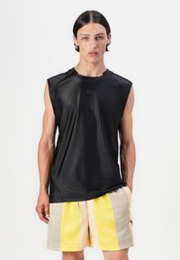 Svart ermeløs treningsskjorte laget av glatt stoff, med en liten logo på brystet. Kombinert med beige og gule stripete shorts.