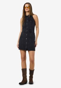 Zwarte denim mouwloze jurk met een knoopsluiting aan de voorkant, een getailleerde taille en twee borstzakken. Bruine kniehoge laarzen maken de look compleet.