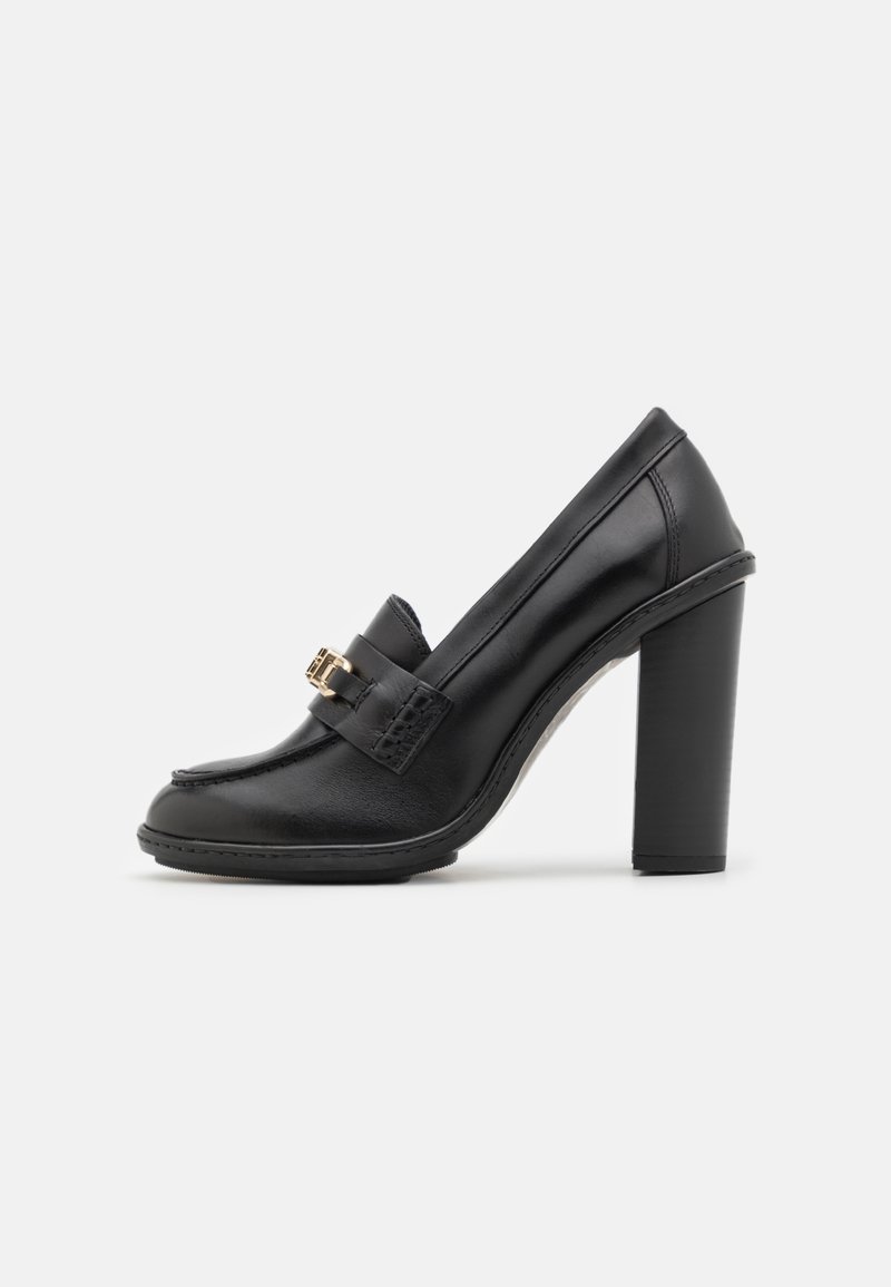 Mocasín negro de cuero con tacón alto y grueso, adornado con un detalle de hebilla dorada en la parte superior, mostrado de lado sobre un fondo blanco.