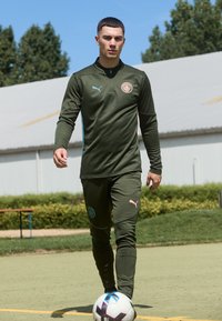 Olívazöld Puma fociedzői öltözet, állítható gallérral, márkajelzéssel és testhezálló kivitelben; a játékos egy focipályán áll, a lába mellett egy focilabdával.