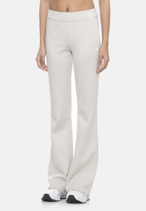 Donna con pantaloni beige chiaro a gamba larga e sneakers bianche, mostrata dalla vita ai piedi su sfondo uniforme.