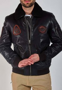 Homme portant une veste en cuir noire avec un col en shearling et des patchs, les mains jointes, portant un pantalon beige, sur fond uni.