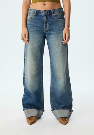 Woman wearing wide-leg faded blue jeans with large cuffs over brown pointed lace-up shoes, standing against a white background.