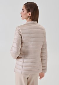 Veste matelassée beige clair avec un motif matelassé horizontal, col côtelé, manches longues et texture lisse. Convient pour les temps plus frais.