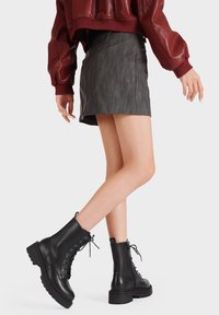 Stivali neri con lacci alla caviglia, suola spessa e punta tonda, abbinati a una minigonna in pelle grigia. L'outfit è completato da una giacca color bordeaux.