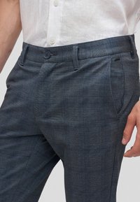 Primo piano di una persona che indossa pantaloni eleganti a quadri grigio scuro con bottone e passanti per cintura, abbinati a una camicia bianca abbottonata.
