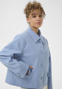 Világoskék gyapjúkeverék dzseki, galléros nyakkal, nagy gombokkal és oldalsó zsebekkel. Lazán illeszkedő fazon és textúrált felület jellemzi.