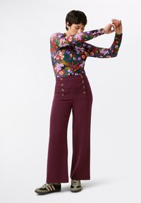 Top floreale a maniche lunghe in verde, rosso e viola; pantaloni a vita alta color bordeaux con sei bottoni decorativi; sneakers verdi e nere.